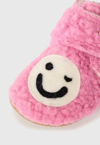 Rosa filttoffel med en strukturerad yta, med en vit smiley-design i mitten och ett kardborreband för säker passform.