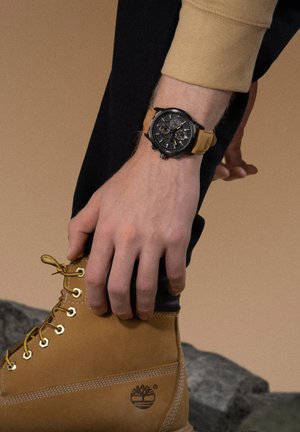 Montre chronographe avec un boîtier noir et un bracelet en cuir tan. Présentant un cadran noir texturé et des accents dorés. Assortie à une botte tan.