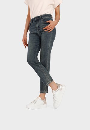 Persona che indossa jeans grigio scuro, sneakers bianche con dettagli metallici e una maglietta a maniche corte di colore chiaro, in piedi davanti a uno sfondo semplice.