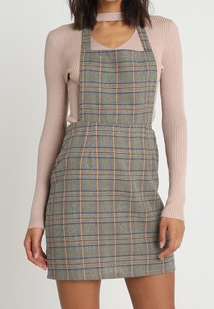 Vrouw draagt een aansluitende grijze geruite tuinbroekjurk over een geribd, lichtroze, lang mouwtopje met een kleine uitsnede bij de halslijn.