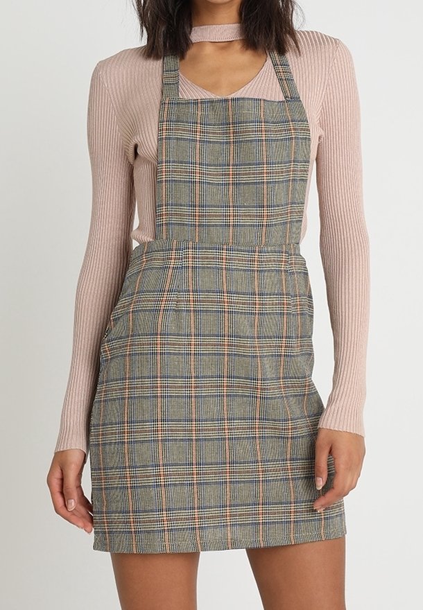 Femme portant une robe salopette ajustée à carreaux gris par-dessus un haut côtelé à manches longues rose clair avec une petite découpe au niveau de l'encolure.