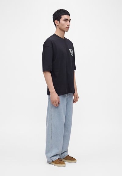 Νέος άνδρας με μαύρο oversize T-shirt με μικρό λογότυπο στο στήθος, ανοιχτό μπλε φαρδιά τζιν και καφέ αθλητικά παπούτσια, στέκεται πλάγια σε λευκό φόντο.