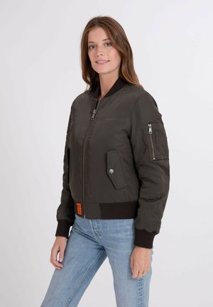 Mujer con una chaqueta bomber verde oscuro con bolsillo con cremallera en la manga y jeans azul claro, de pie frente a un fondo blanco liso.