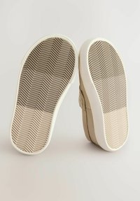 Beige halkskor med ett texturerat tygöverdrag och gummisålar. Sålarna har ett fiskbensmönster för bättre grepp. Vit kant runt basen.