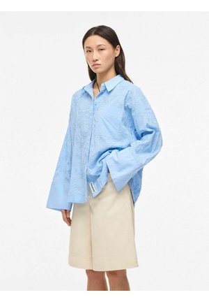 Giovane donna che indossa una camicia oversize azzurro chiaro con bottoni e pantaloncini beige fino al ginocchio su sfondo bianco.