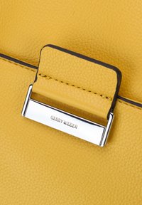 Szczegół żółtej toczonej skórzanej torebki na ramię z metalowym zapięciem w kolorze srebrnym, na którym wygrawerowano "GERRY WEBER". Gładkie krawędzie z ciemnym wykończeniem.