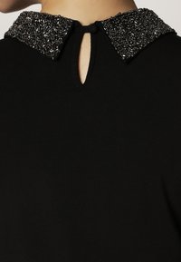 Svart plagg med en närbild av en pärlad krage som har svarta och mörk-silverfärgade pärlor samt en liten central nyckelhålsutskärning.