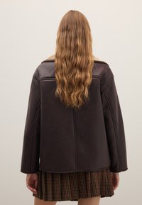 Braune Lederjacke mit Schafsfellkragen, weiten Ärmeln, sichtbaren Nähten und einem leicht übergroßen Schnitt, getragen über einem karierten Rock.