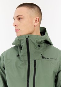 Jonge man met kort geschoren haar in een groene gevoerde jas met een zwarte rits en de tekst "PROJECT PVRE GREEN" op de borst.