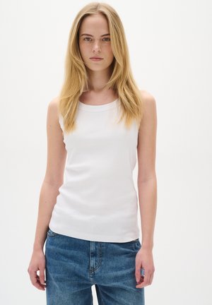 Junge Frau mit glattem blondem Haar, die ein ärmelloses weißes Top und blaue Jeans trägt, vor einem schlichten hellen Hintergrund stehend.