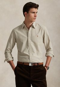 Junger Mann trägt beige kariertes Hemd mit hochgekrempelten Ärmeln, braunen Gürtel, dunkle Hose und Armbanduhr, Hände in den Taschen, schaut nach rechts.