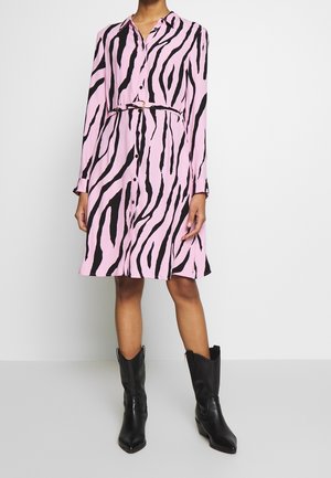 Mujer con un vestido de largo hasta la rodilla, estampado de cebra en rosa y negro, con mangas largas y cinturón, combinado con botas de cuero negras hasta la mitad de la pantorrilla.