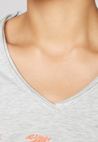 Šedé, lehké tričko s výstřihem do V. Látka vypadá měkce a má mírný lesk; obsahuje oranžový potisk v oblasti hrudníku.