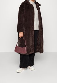 Bruine jas van imitatiebont met kraag, knoopsluiting, grijze trui, zwarte corduroy broek en een kleine bordeauxrode handtas met krokodillenprint.