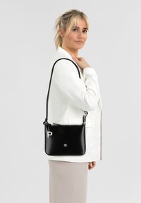 Schwarze Leder-Umhängetasche mit silbernen Beschlägen, ausgestattet mit einem Reißverschluss und einem auffälligen Buchstaben "P" Akzent. Glatte Textur und rechteckige Form.