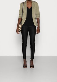 Blazer vert olive, haut noir à bordure festonnée, pantalon skinny noir et talons noirs à brides, assortis à un sac bandoulière rayé noir et beige.