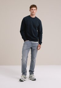 Jonge man in een donkere sweatshirt en lichte spijkerbroek staat zelfverzekerd op een neutrale achtergrond, gekleed in sportieve schoenen.