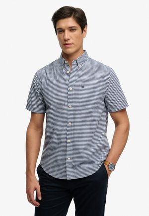 Młody mężczyzna w krótkim, niebiesko-białym koszuli w kratę zapinanej na guziki i ciemnych spodniach, z zegarkiem na lewym nadgarstku.