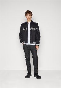 Svart varsityjacka med vit text, vit t-shirt, mörka byxor och svarta stövlar. Jackan har ribbade cuffs och nederkant.