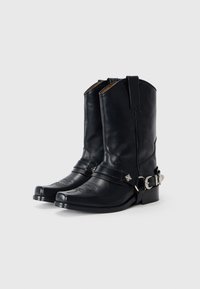 Botas de vaquero de cuero negro con punta afilada, costuras detalladas, correa decorativa con herrajes plateados y tacón cuadrado.