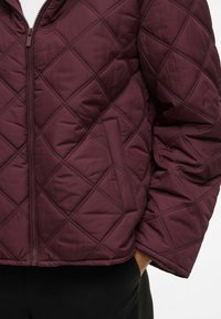 Burgundy quilted jacket featuring a zip closure, diamond pattern, and side pockets with a smooth finish.