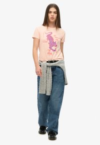 Camiseta de manga corta de color rosa claro con un diseño gráfico en morado. Combinada con vaqueros holgados azules y un suéter gris atado a la cintura.
