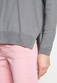 Maglione grigio lavorato a maglia con orlo a coste, caratterizzato da uno spacco laterale. Abbinato a pantaloni rosa chiaro, evidenziando il contrasto tra i colori e le texture.