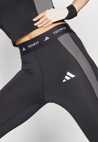 Sorte kompresjonsleggings med et grått sidepanel, med en hvit "TechFit"-livlinning og tre hvite striper logo på låret.