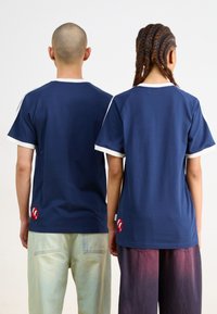 Marine bomull t-skjorter med hvite detaljer og korte raglanermer, med en liten rød logo på nederste venstre side. Bakvisning vist.