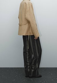 Vaaleanbeige, pellavamainen pitkähiha shirt, jossa on vyö vyötäröllä, yhdistettynä musta-harmaisiin pystysuoriin, löysiin housuihin.