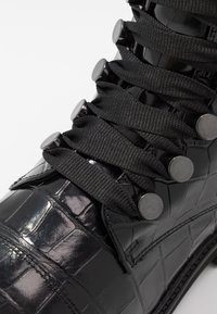 Czarne buty z sztucznej skóry krokodyla z okrągłymi, czarnymi metalowymi oczkami, płaskimi czarnymi sznurówkami oraz teksturowaną powierzchnią z połyskiem.