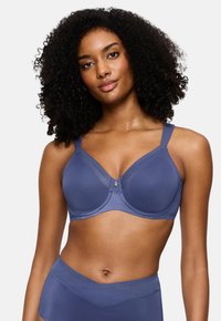 Ženska s kodrasto pričesko, oblečena v modro prekrivalo in hlačne hlačke z visokim pasom, pozira na enobarvni beli podlagi.