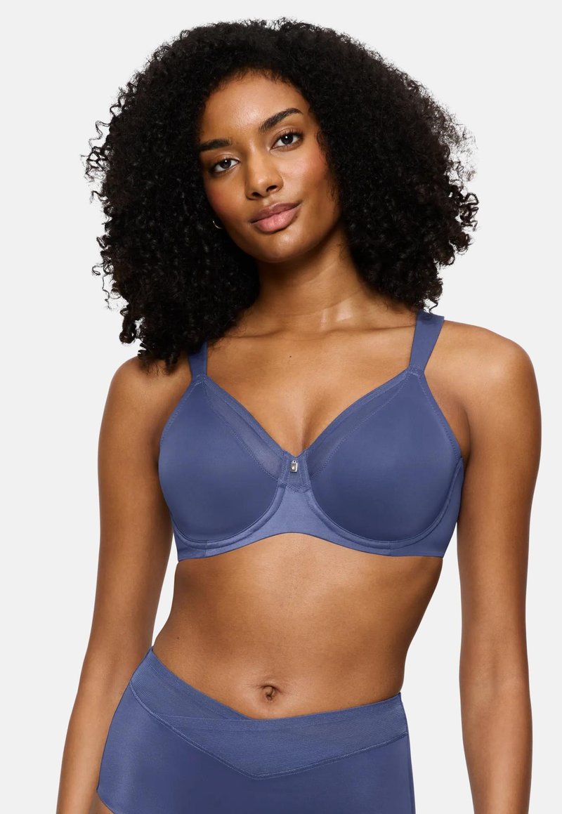 Ženska s kodrasto pričesko, oblečena v modro prekrivalo in hlačne hlačke z visokim pasom, pozira na enobarvni beli podlagi.