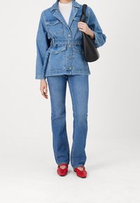 Giubbotto di denim con lavaggio chiaro, bottoni frontali e due tasche sul petto, abbinato a jeans coordinati e scarpe basse rosse, mentre tiene una borsa nera.