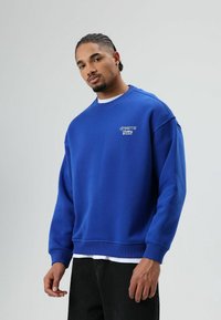 Sweat-shirt bleu à col rond en tissu doux, avec une ceinture et des poignets côtelés, orné d'un texte brodé sur la poitrine. Porté par-dessus une chemise blanche.