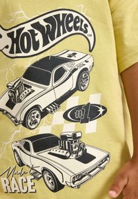 Keltainen T-paita, jossa on mustia graafisia kuvastoja kahdesta kilpa-autosta, joissa on yksityiskohtaiset moottorit ja renkaat, sekä "Hot Wheels" -logo ylhäällä.
