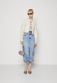 Veste cropped en cuir crème, t-shirt graphique gris, jupe en denim bleu clair avec poches, mini sac à main rose, ballerines marron à bout pointu, lunettes de soleil noires.