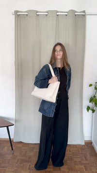 Geantă de tote din pânză albă, cu o formă structurată și un umăr lat, asortată cu o jachetă din denim și pantaloni negri, într-un cadru minimalist.