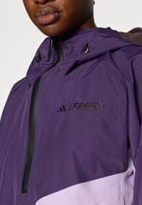 Ljubičasta vodootporna jakna s crnim patentnim zatvaračem, podesivom kapuljačom i izvezenim logotipom "TERREX" u tamnoljubičastoj boji na prsima.
