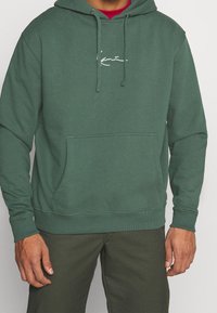 Sudadera verde con capucha de cordón, bolsillo frontal y logo blanco. Tejido suave con un ajuste relajado y puños y dobladillo acanalados.