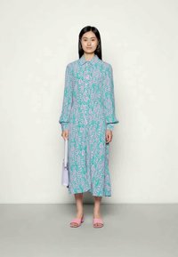 Vestido longo com um padrão ondulado em verde e roxo, frente com botões, mangas longas, usado com sandálias rosa e uma bolsa lilás clara.