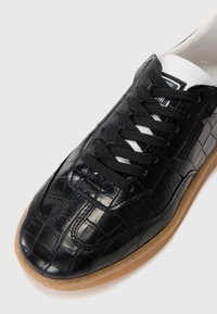 Sort sneakers lavet af struktureret, blankt materiale med et krokodillemønster, der har sorte snørebånd og en sål af gummi.