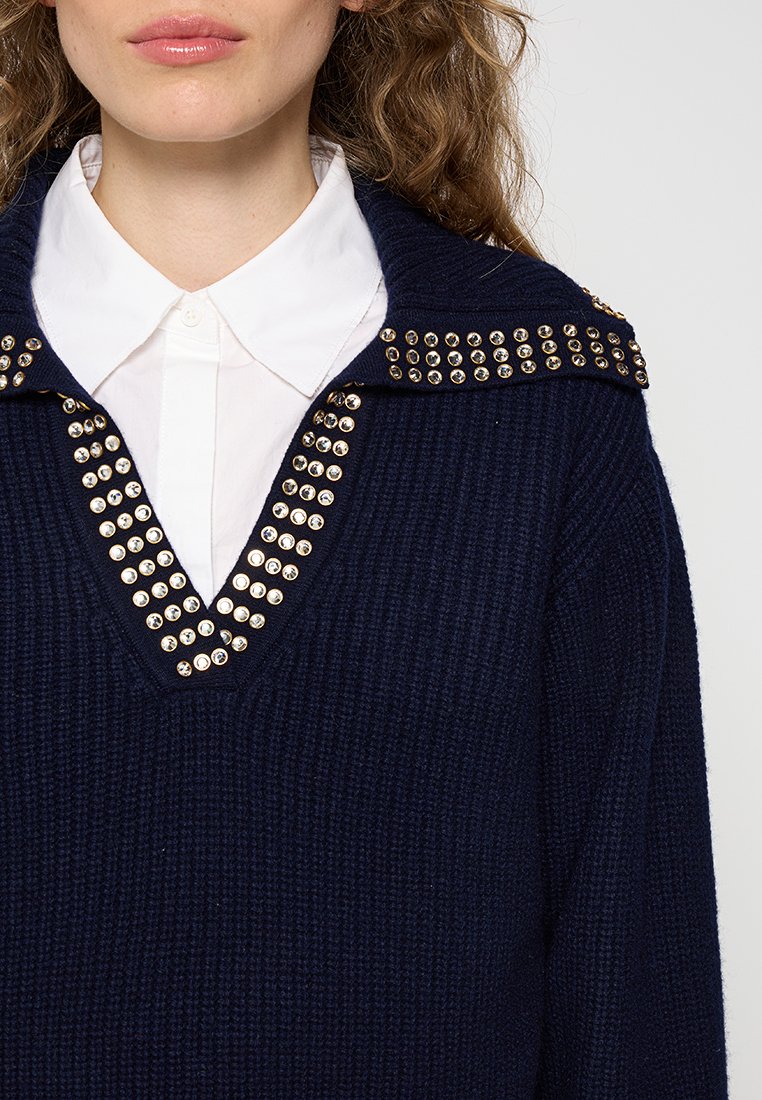Pull en tricot navy avec un col en V et des accents de col cloutés dorés. Présente un tissu texturé et une chemise blanche contrastante en dessous.