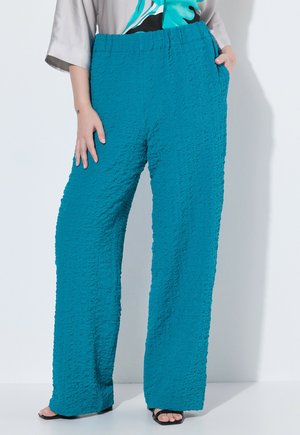 Donna che indossa pantaloni larghi turchesi con texture e vita elastica, abbinati a un top a fantasia e sandali neri open toe.