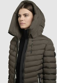 Pūtīga tumši olīvzaļā jakas, ar lielu kapuci, melnu rāvējslēdzēju un sānu rāvējslēdzēja kabatām. Iespēja ar gludu tekstūru un quilted dizainu.