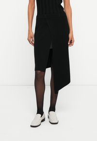 Melnā adīta svārki ar augstu šķēlumu, ar teksturētu apdari un platu jostasvietu. Kombinēti ar baltām plakanām kurpēm un caurspīdīgām melnām zeķbiksēm.