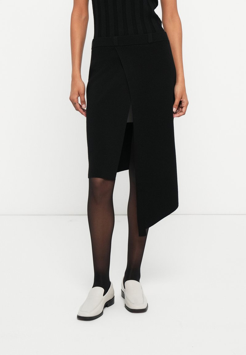 Melnā adīta svārki ar augstu šķēlumu, ar teksturētu apdari un platu jostasvietu. Kombinēti ar baltām plakanām kurpēm un caurspīdīgām melnām zeķbiksēm.