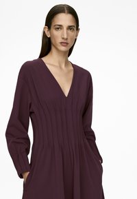 V-ausschnitt lila Kleid mit langen Ärmeln, seitlichen Taschen und vertikalen Paspelierungen. Aus glattem, leichtem Stoff gefertigt.