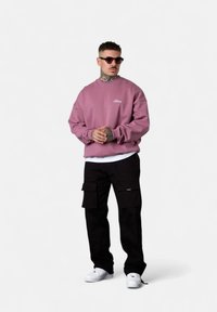 Oversized pink sweatshirt with white logo, layered over a white shirt, paired with black cargo pants and white sneakers, worn with sunglasses.