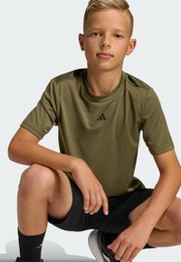 Olivovo zelené športové tričko s krátkym rukávom a okrúhlym výstrihom s čiernym logom Adidas, doplnené čiernymi šortkami a čiernymi ponožkami.